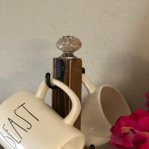 May include: A wooden mug rack with a white ceramic knob on top. The rack has five hooks and is holding four white mugs. One mug has the word "Beast" written on it in black.