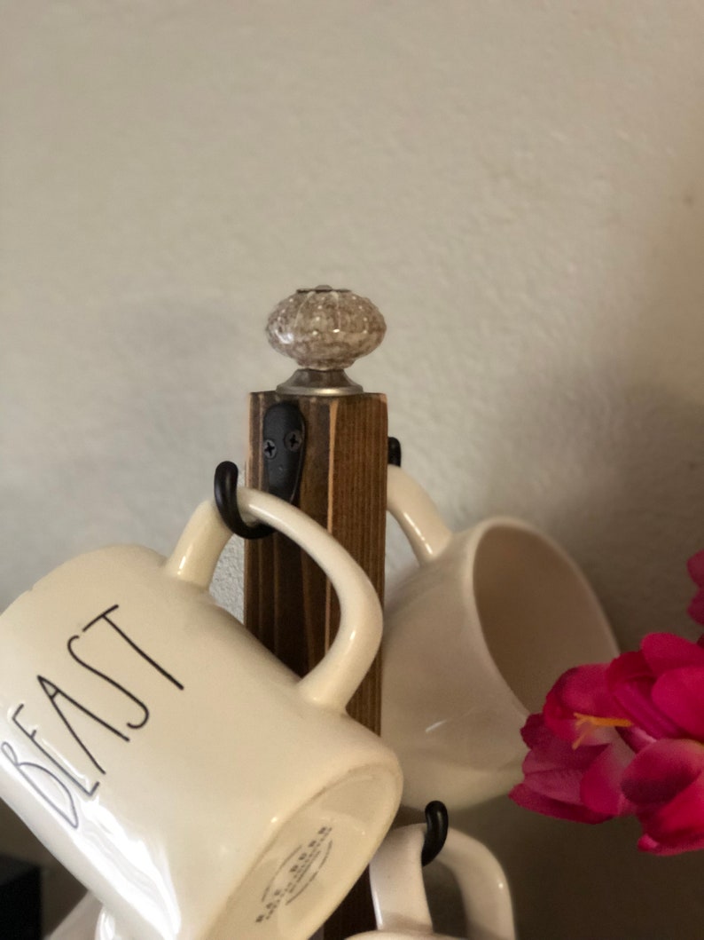 May include: A wooden mug rack with a white ceramic knob on top. The rack has five hooks and is holding four white mugs. One mug has the word "Beast" written on it in black.