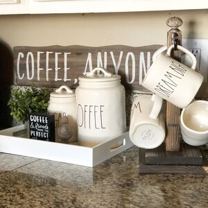 May include: A wooden mug rack with four white ceramic mugs hanging from it. The mugs have black lettering that reads "Dream Big" and "Mom". The rack is sitting on a granite countertop. There are also three white ceramic canisters with black lettering that reads "Coffee", "Sugar", and "Coffee". A small black sign with white lettering that reads "Coffee & Friends make the perfect day" is sitting on a white tray.