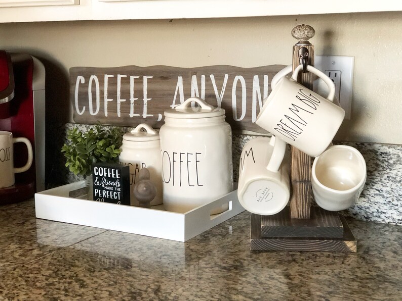 May include: A wooden mug rack with four white ceramic mugs hanging from it. The mugs have black lettering that reads "Dream Big" and "Mom". The rack is sitting on a granite countertop. There are also three white ceramic canisters with black lettering that reads "Coffee", "Sugar", and "Coffee". A small black sign with white lettering that reads "Coffee & Friends make the perfect day" is sitting on a white tray.