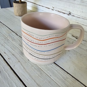 May include: A light pink ceramic mug featuring a handle and horizontal wavy lines in orange, blue, and brown. The mug displays the text: I HOPE IN HALLWAYS AND PARKING LOTS YOU CAN TAKE A MOMENT TO BREATHE, TO LOOSEN YOUR SHOULDERS AND KNOW IT IS OKAY TO NOT KNOW EVERYTHING. MHN.