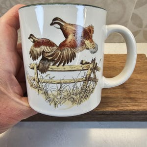 May include: White ceramic mug featuring a detailed illustration of two birds in flight over a wooden fence and tall grass. The mug has a green rim and a curved handle, perfect for enjoying hot beverages.