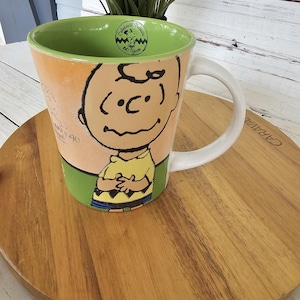 May include: A ceramic mug featuring Charlie Brown from the Peanuts comic strip. The mug has a white handle and a green interior. The exterior is decorated with a cartoon image of Charlie Brown in a yellow shirt and black shorts. The mug also has the text "Celebrate Peanuts 60 Years".