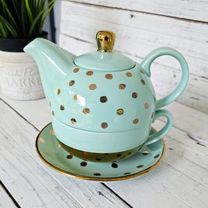May include: A mint green ceramic tea set with gold polka dots. The set includes a teapot with a gold lid, a teacup, and a saucer. The saucer and the rim of the teapot are trimmed with gold.