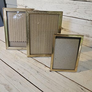 May include: Three gold-colored picture frames of varying sizes. The frames have a rectangular shape with a decorative border. The frames are empty, with cardboard backing visible. The frames are arranged on a white wooden surface.