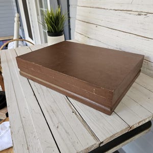 May include: A rectangular, brown wooden box with a slightly distressed finish. The box is closed and sits on a white, weathered wooden surface. The box appears to be a storage container or decorative item.