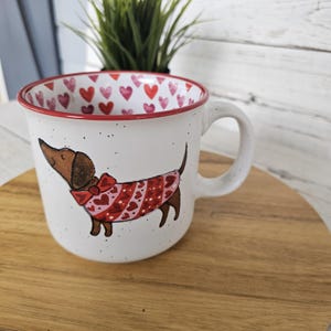 May include: White ceramic mug with a red rim and handle, featuring a dachshund wearing a red sweater with heart patterns. The inside of the mug is decorated with red and pink hearts. The mug is speckled with small black dots.