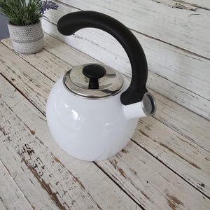 May include: White enamel tea kettle with a black handle and a silver lid. The kettle is sitting on a white wooden surface.