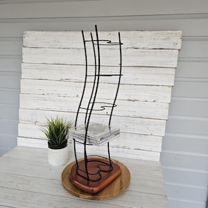 May include: A black metal CD rack with a wavy design. The rack has several shelves and sits on a wooden base. A small potted plant is to the left of the rack. The background is a white wooden wall.