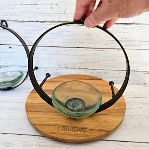 May include: A decorative wall sconce featuring a round, wooden base with the word "CARAVAN" engraved on it. A black metal ring holds a clear, green-tinted glass bowl. The sconce is designed for displaying small items.