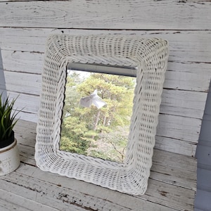May include: White wicker framed mirror with a woven design. The mirror reflects a view of trees and a white object.