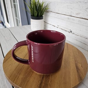 May include: A burgundy ceramic coffee mug with the Starbucks logo in white. The mug sits on a round wooden coaster. A small potted plant is visible in the background.