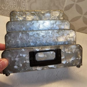 May include: A galvanized metal desk organizer with four tiered compartments. The organizer has a rectangular black label holder on the front and rests on small metal ball feet. The metal has a mottled, textured appearance.