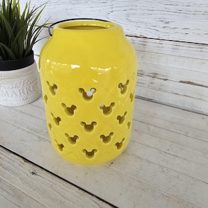 May include: A bright yellow ceramic lantern with Mickey Mouse-shaped cutouts. The lantern has a black handle and sits on a white wooden surface. A small potted plant is visible in the background.