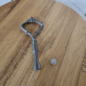 May include: A silver-colored metal handle with an ornate, decorative top and a small, round silver-colored screw. The handle is designed with a curved shape and a textured grip. The items are on a wooden surface.