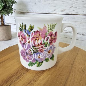 May include: A white ceramic mug featuring a colorful floral arrangement of pink, purple, and blue flowers with green leaves. The text "Hung Over" is printed in a white cursive script font across the center of the floral design on the mug.