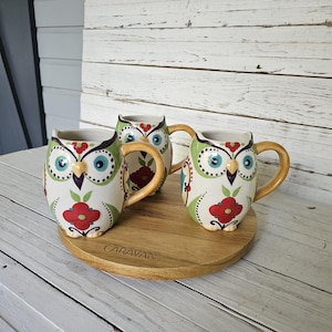 May include: Three ceramic owl mugs with colorful floral designs and tan handles sit on a wooden serving tray. The mugs feature a white base with green, red, and blue accents. The tray has the word "CARAVAN" engraved on it.