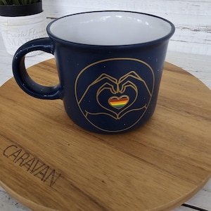 May include: A navy blue ceramic mug with a white interior. The mug features a gold outline of hands forming a heart, with a rainbow heart in the center. The mug is on a wooden serving board.