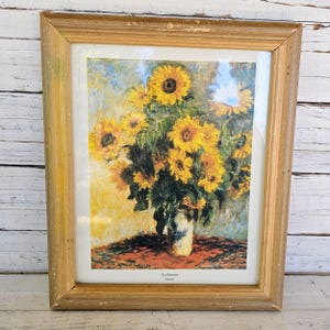 May include: Framed print of sunflowers in a vase. The artwork features a bouquet of yellow sunflowers with green leaves in a blue and white vase. The frame is made of light brown wood.