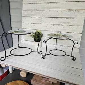 May include: Two-tiered black metal serving stands with decorative scrollwork. Each stand has a round base and a circular top with a plate featuring a green leaf design. A small potted plant sits between the stands.