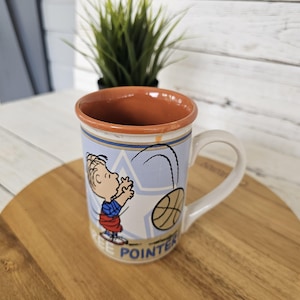 May include: A white ceramic mug with a cartoon illustration of a boy playing basketball. The mug has a light blue background with a basketball design and the words "FREE POINTER". The interior is brown.