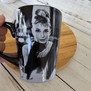 May include: Black coffee mug featuring a black and white image of Audrey Hepburn. The mug has a black handle and a glossy finish. The image shows Hepburn wearing a pearl necklace and a black dress. The mug is on a wooden surface.