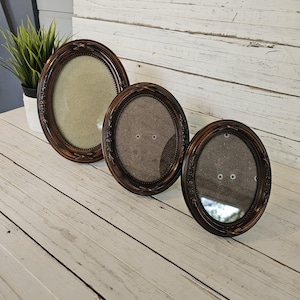 May include: Three oval-shaped picture frames in varying sizes. The frames are a dark brown with ornate detailing around the edges. The frames have a glass front and a backing material. The frames are displayed on a white wooden surface.