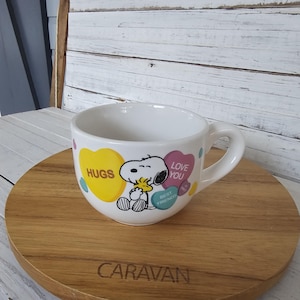 May include: White ceramic mug featuring Snoopy and Woodstock hugging. The mug has yellow, pink, and blue heart-shaped designs with the words "HUGS", "LOVE YOU", and "BEST FRIENDS". The mug sits on a wooden serving tray.