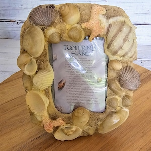 May include: A decorative photo frame with a sandy, seashell-encrusted border. The frame holds a printed poem titled "Footprints in the Sand." The frame is beige and brown, with various seashells and a starfish accenting the border.