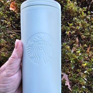May include: A light blue Starbucks tumbler with a lid, held by a hand. The Starbucks logo is embossed on the front. The tumbler is cylindrical and appears to be made of metal or a similar material. The background is a green bush.