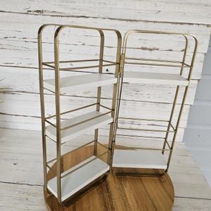 May include: Two decorative wall shelves. Each shelf has a gold-colored metal frame with three white shelves. The shelves are rectangular and have a minimalist design. The shelves are placed on a wooden surface.