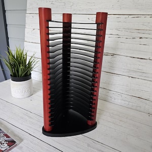 May include: A red and black CD rack with a fan-like design. The rack has a wooden frame with a curved base and top, and black dividers to hold CDs. The rack is on a white wooden surface.