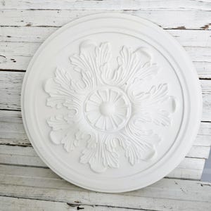 May include: A white, circular ceiling medallion with an ornate, floral design. The medallion features a central rosette surrounded by detailed, stylized leaves and a raised outer edge. The decorative element is made of a solid, opaque material.