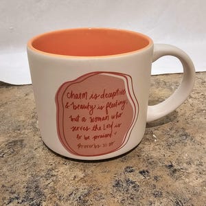 May include: A cream-colored ceramic mug with a peach-colored interior and handle. The front features a pink, organic-shaped graphic with the text: charm is deceptive & beauty is fleeting; but a woman who serves the Lord is to be praised. Proverbs 31:30.