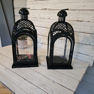 May include: Two black metal lanterns with arched glass panels and decorative lattice cutouts at the top. One lantern contains a small potted plant and thin, glowing red string lights, while the other lantern is empty with a clear glass interior.