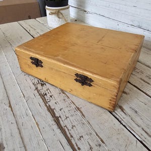 May include: A wooden box with two metal clasps. The box is light brown and has a smooth surface. The box is sitting on a white wooden surface.