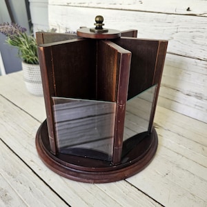 May include: A rotating dark brown wooden carousel organizer featuring multiple vertical compartments with clear glass panels on the front. The unit has a circular base and a small brass-colored knob at the top center for easy rotation.