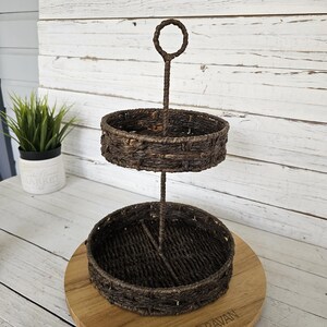 May include: A two-tiered brown woven serving tray with a handle. The tray has two circular baskets and a central support. The tray is on a wooden surface with a plant in the background.