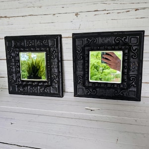 May include: Two square, black decorative mirrors with ornate, embossed frames. Each mirror has a small, square reflective surface. One mirror reflects a plant, the other reflects a hand holding a phone. The mirrors are displayed on a white, wooden surface.