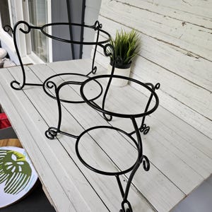 May include: Three black metal plant stands with circular platforms and decorative scrollwork. The stands have three legs and are designed to hold potted plants. The stands are on a white wooden surface.