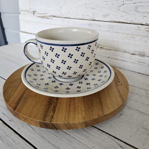 May include: A white ceramic teacup and saucer set with a blue floral pattern. The cup has a handle and sits on a matching saucer. The set is placed on a wooden serving board.