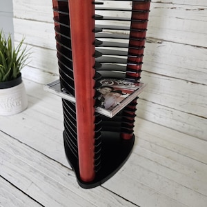 May include: A black and red CD storage tower with multiple slots for discs. The tower has a red central support and black shelves. A CD case is visible on one of the shelves. The tower is on a white wooden surface.