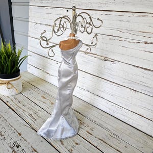 May include: A silver dress form jewelry holder with a metal top for hanging earrings and necklaces. The dress form has a satin-like finish and a mermaid-style skirt. The jewelry holder is displayed on a white wooden surface.