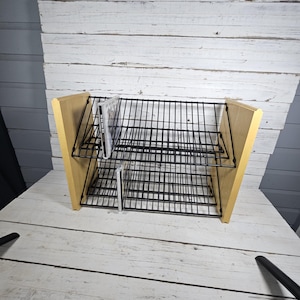May include: A two-tier media storage rack featuring light-toned wooden side panels and a black wire grid frame. The rack is designed with an angled shelf structure to hold items upright, and it currently contains two slim plastic cases.