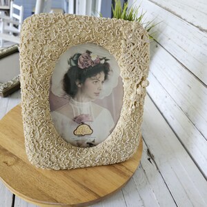 May include: Cream-colored beaded picture frame with an oval opening. The frame features a portrait of a woman wearing a floral headpiece. The frame is on a wooden surface. The frame is labeled "ANNO FRAMES".