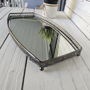 May include: An antique-style mirrored tray with a decorative metal frame. The tray has a rectangular shape with rounded ends and sits on small ball feet. The frame features an intricate design and a dark, aged finish. A small potted plant is visible in the background.
