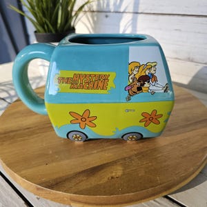 May include: A turquoise ceramic mug shaped like the Mystery Machine van from Scooby-Doo. The mug features the van's design, including the text "The Mystery Machine" and cartoon characters. The mug has a large, rounded handle and is sitting on a wooden surface.