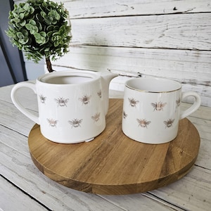 May include: A white ceramic teapot and a matching mug, both decorated with a repeating pattern of small, metallic gold-toned bees. The mug features a thin gold rim along the top edge. Both items have a smooth, glossy finish and simple handles.