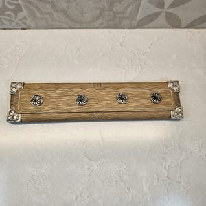 May include: A rectangular wooden wall-mounted key holder with four metal hooks. The wood has a textured grain pattern and is trimmed with decorative metal corners. The hooks are circular with an ornate design.