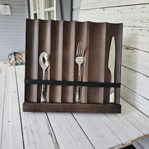 May include: A wooden cutlery holder with a wavy design, holding a spoon, fork, and knife. The holder is dark brown with a black band to secure the utensils. The spoon, fork, and knife are made of shiny metal.